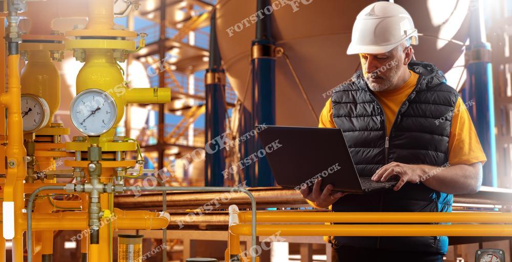 Мужчина в защитной каске и жилете работает с ноутбуком рядом с газовыми трубами и манометрами на промышленной площадке. Оборудование окрашено в желтый цвет, а обстановка подчеркивает серьезность и технологичность контроля параметров в сфере энергетики или газораспределения.